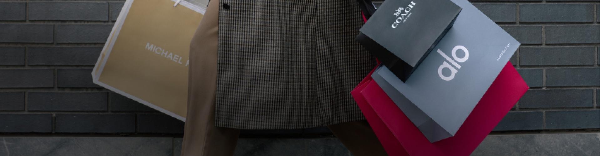 Shopper holding Michael Kors, Coach and Alo bags against a brick wall