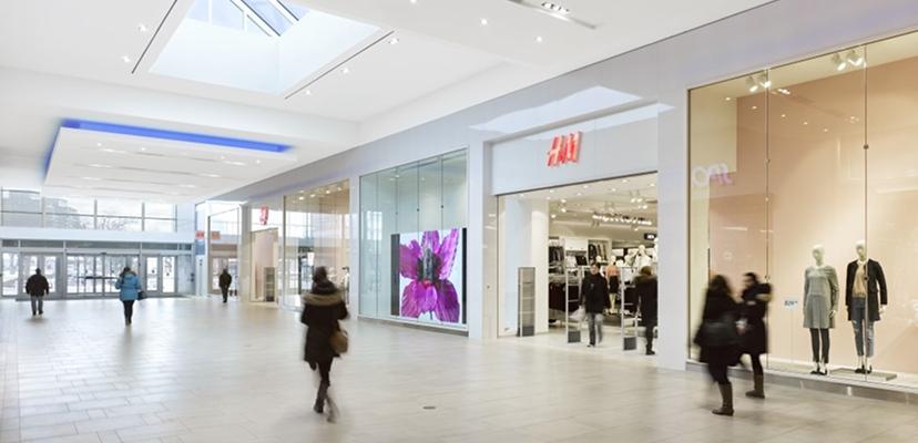 Shoppers in a sunlit mall walkway near H&M with a flower display screen