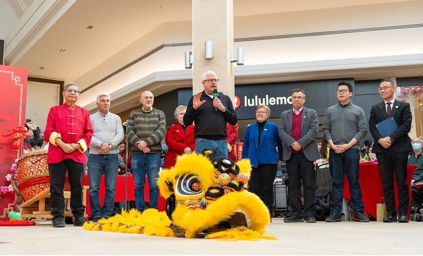Man speaking into a mic beside a lion dance costume at a mall event