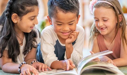 Three smiling children lying down reading a book together