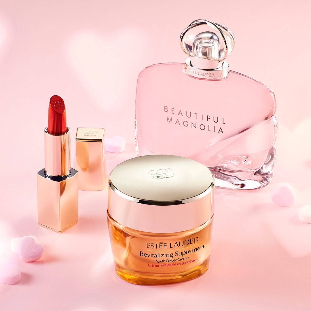 Estee Lauder lipstick, face cream, and perfume are posed on a light pink background.