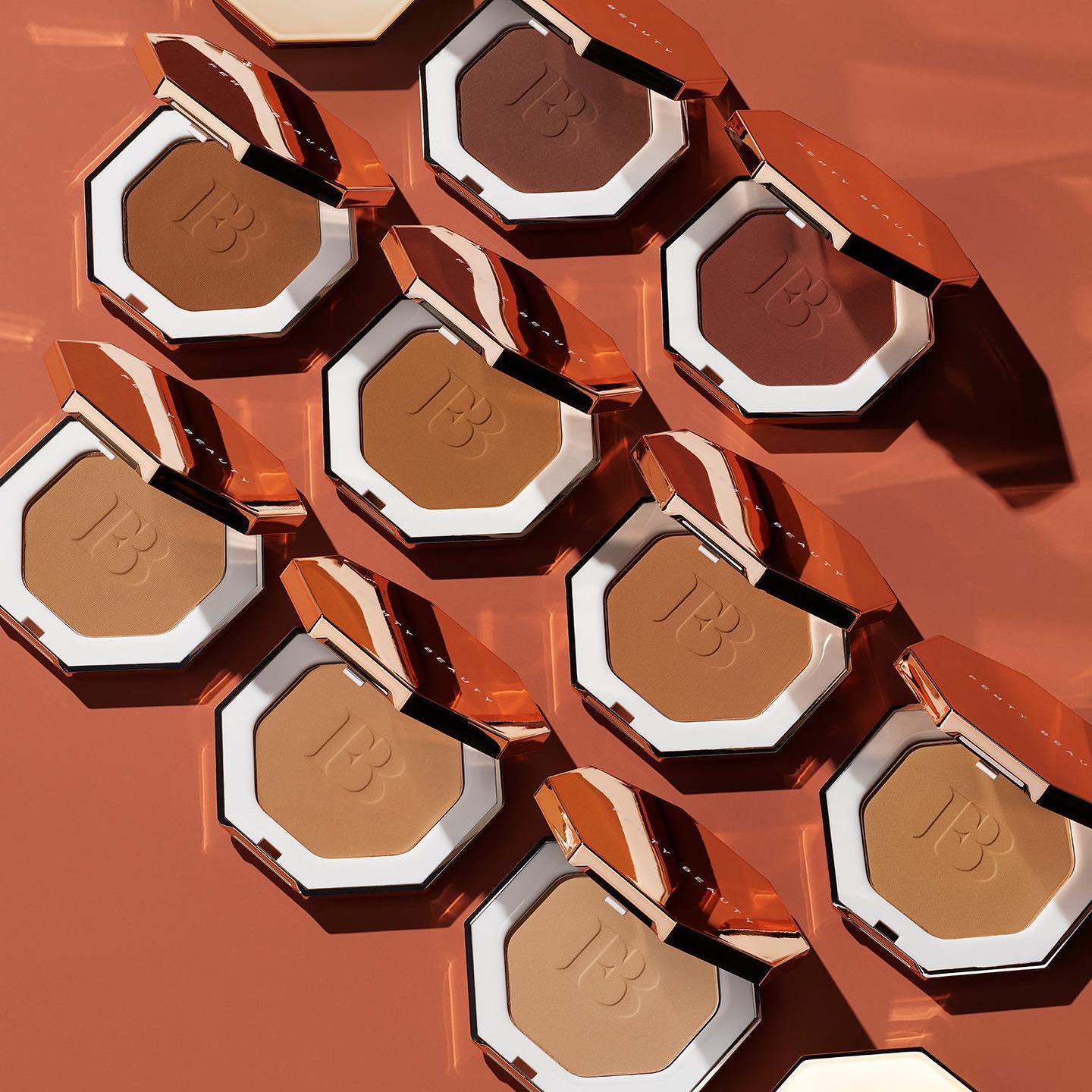 On a rusty orange beackground, various skin shades of Fenty Beauty bronzer are showcased.