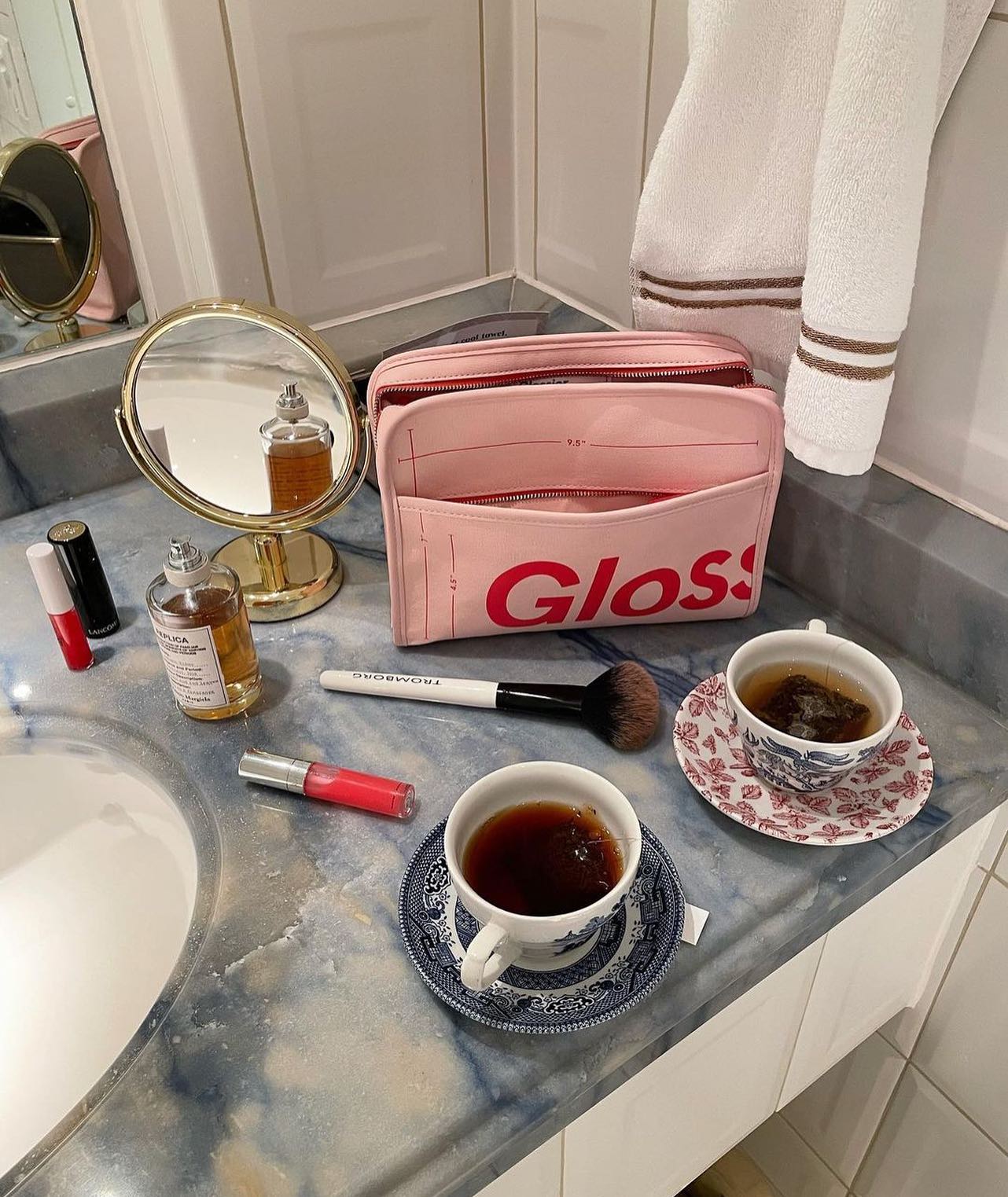A Glossier make up bag is positioned on a bathroom counter top alongside cups of tea and other Glossier products.