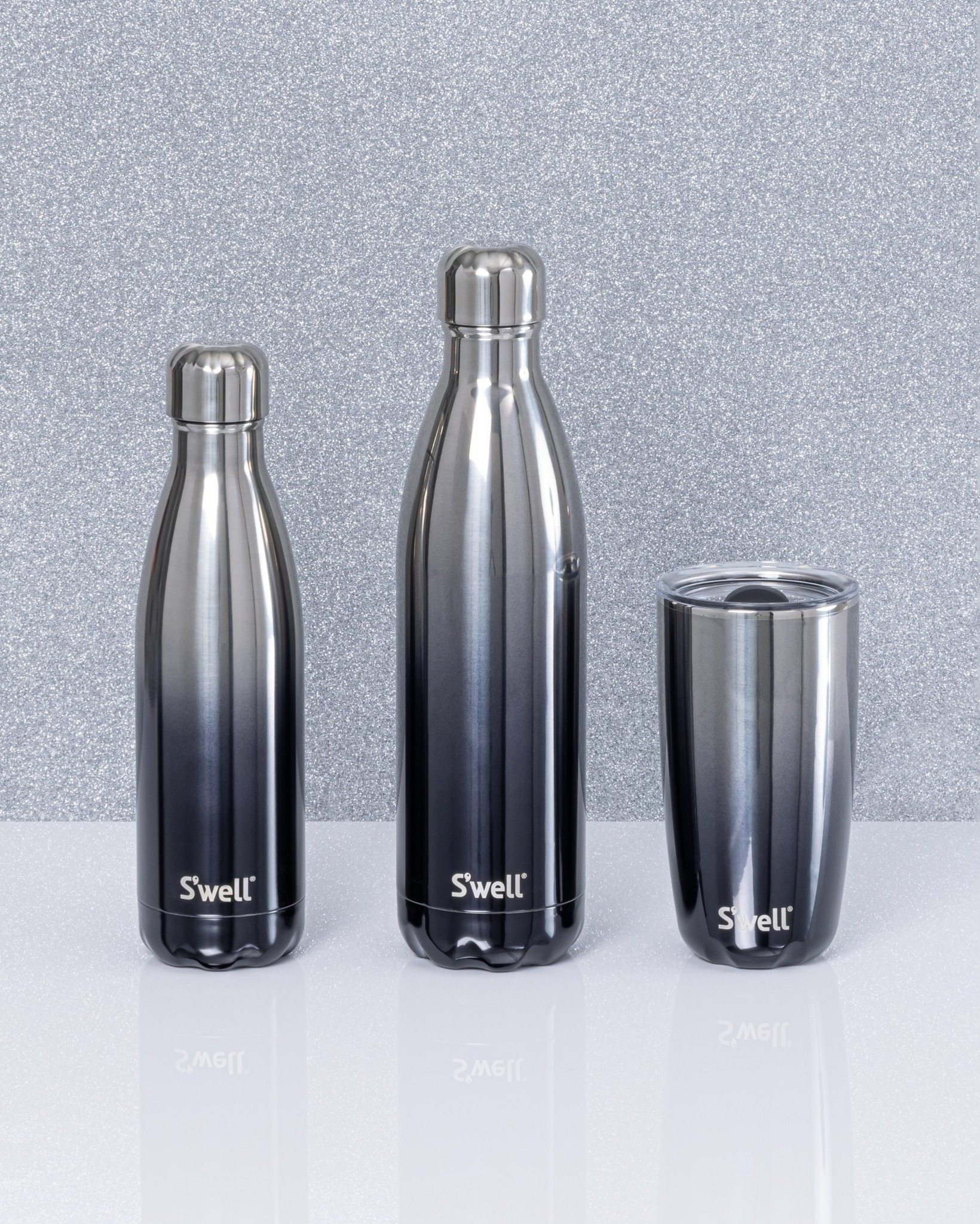 Three various sizes of S'Well water bottles in the same black to silver ombre colour are positioned on a countertop.