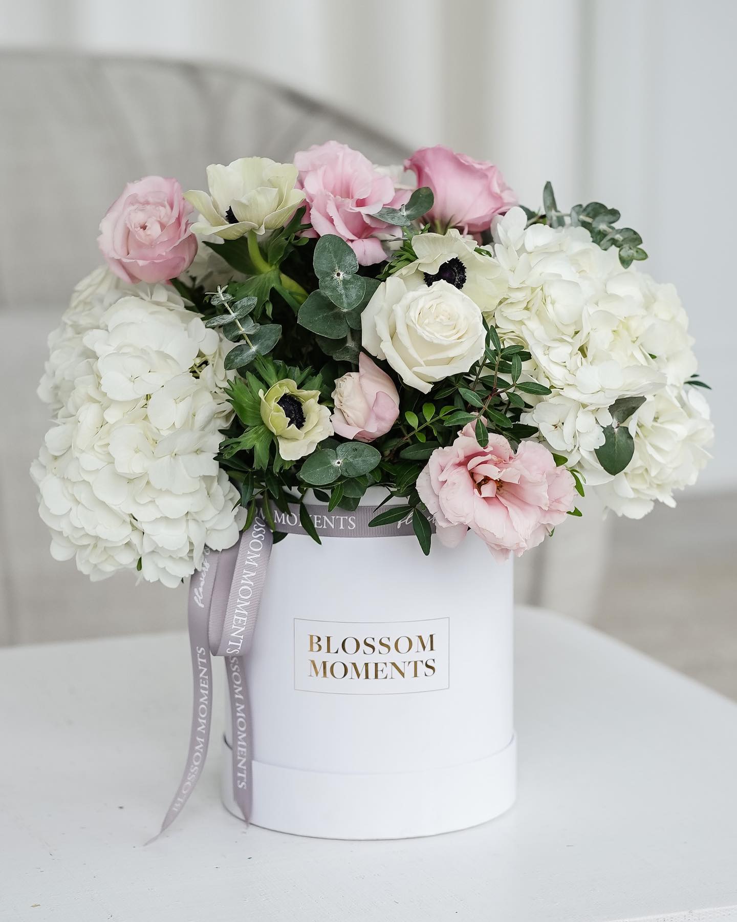 A light pink and white flower arrangement in a container that says Blossom Moments.