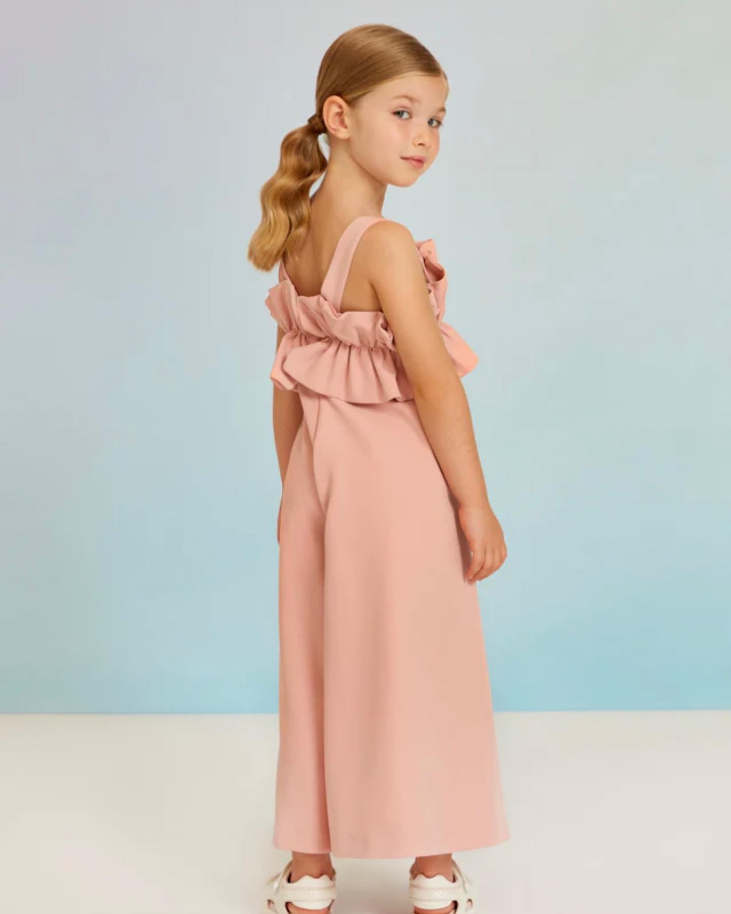 A young girl looks over her shoulder while wearing a light pink jumpsuit from Hunter & Shaye.
