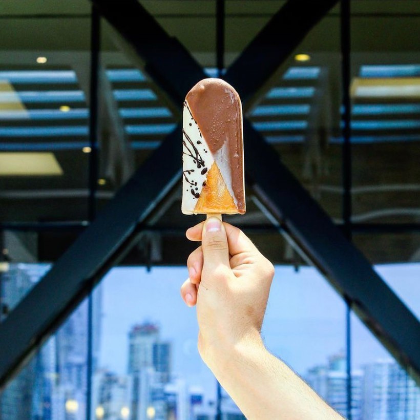 A person's hand is holding an ice cream bar on a stick with a background showing a cityscape.
