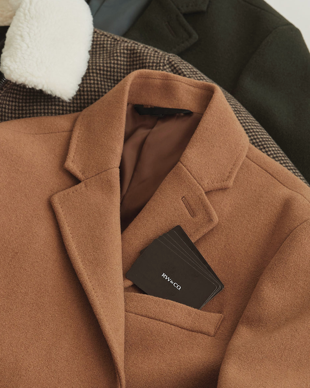 An RW&Co. camel coloured coat is featured laying on top of other clothing.