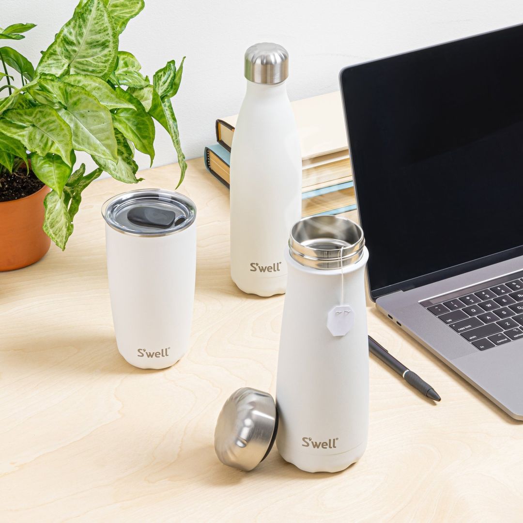 image of three swell products on a light-coloured desk: in the foreground is an open tumbler with a visible tea bag hanging out of it, to its right is a coffee tumbler and in the back is a water bottle. All are white with silver accents. Also in the image is a laptop, some books, a plant and a pen.