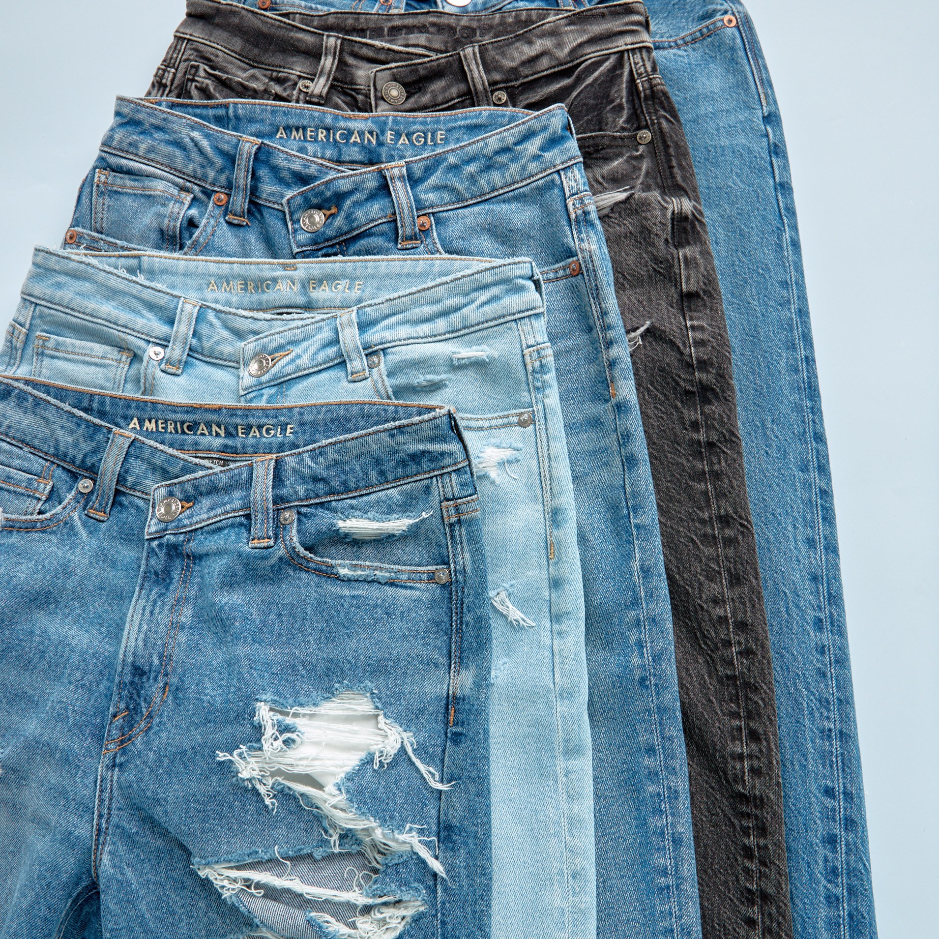 Image of five pairs of jeans layered neatly on top of each other on a slight angle. The first three pairs (starting from bottom) are in various shades of blue and show some distressing, the fourth pair is black and the last pair is blue.