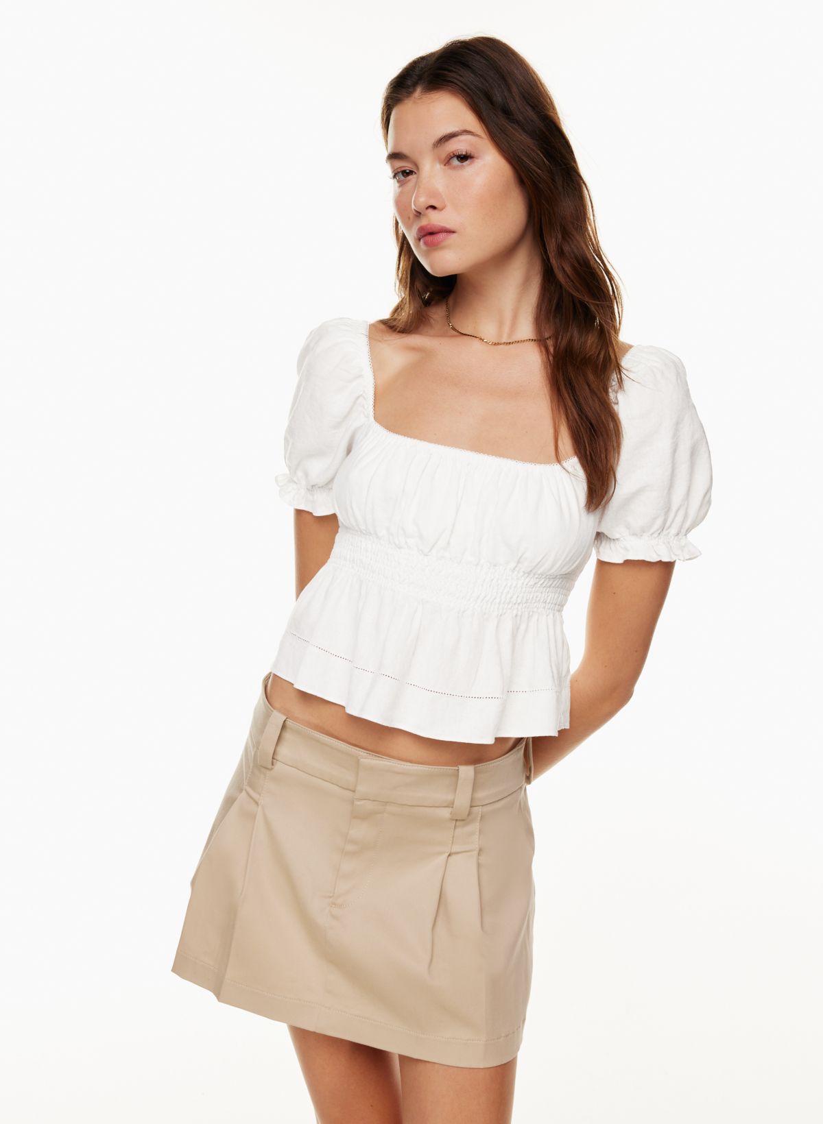 image of a female model with long brown hair wearing a peasant-style white top with a low, straight neckline and slightly puffy short sleeves. The top flares out slightly in the bottom. She pairs the shirt with a khaki-coloured mini skirt.