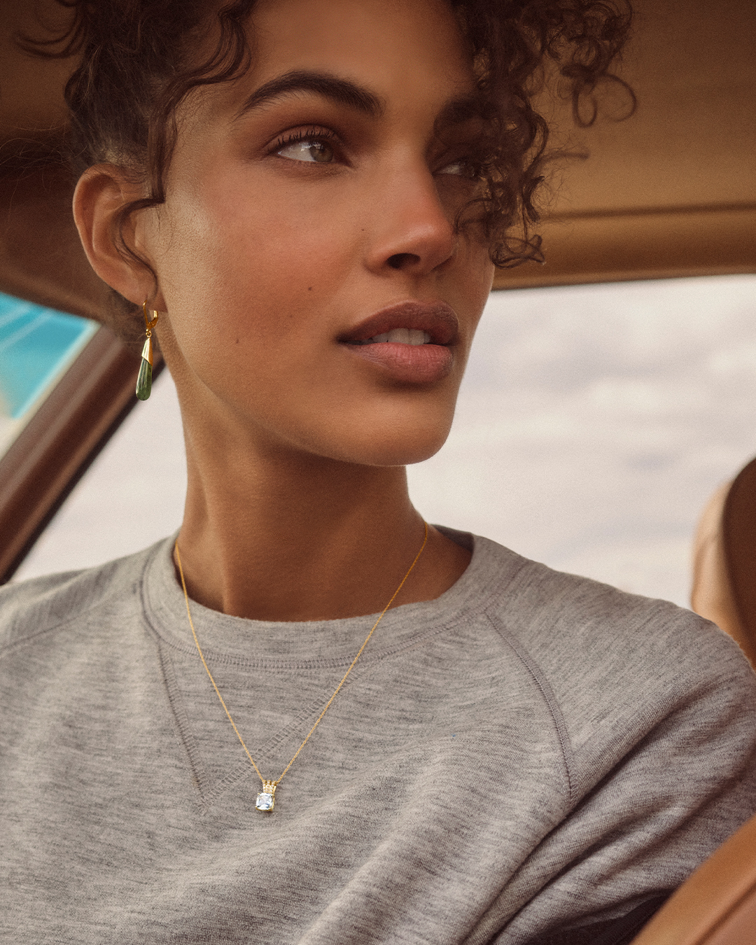 A woman sits in a car and looks off to the distance while showcasing earrings and a necklace from Peoples Jewellers.