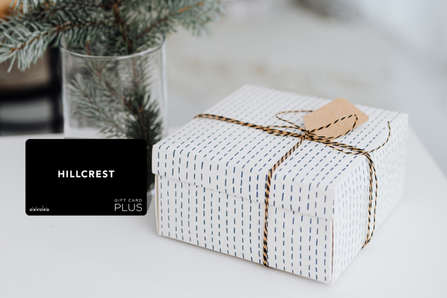 promotional image featuring a black hillcrest gift card. beside the gift card is a present wrapped in white wrapping paper and behind the gift card is a vase with pine tree branches.